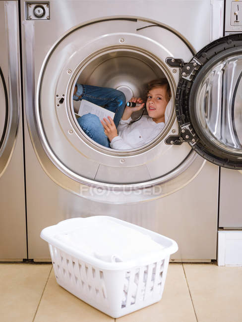 Junge im Grundalter mit Taschenlampe liegt in Waschmaschine mit Buch. — Stockfoto