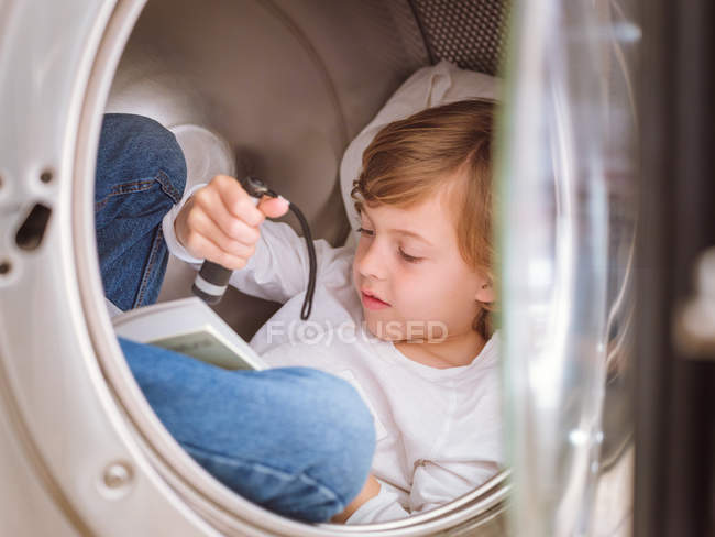 Menino da idade elementar com lanterna deitado dentro da máquina de lavar roupa e livro de leitura . — Fotografia de Stock