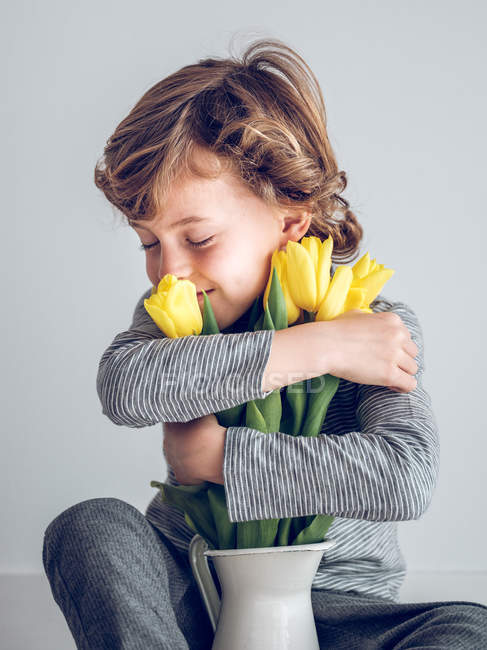 Старшеклассник с закрытыми глазами сидит и обнимает кучу желтых тюльпанов на сером фоне . — стоковое фото