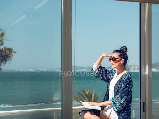 Junge Frau entspannt mit Buch und Blick auf den Balkon am Meer. — Stockfoto