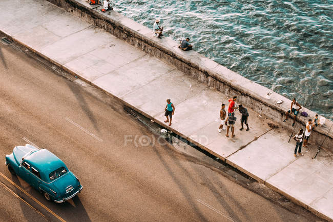 LA HABANA, CUBA - 1 DE MAYO DE 2018: personas descansando en el paseo marítimo pavimentado con agua corriente y autos retro conduciendo por carretera, Cuba - foto de stock