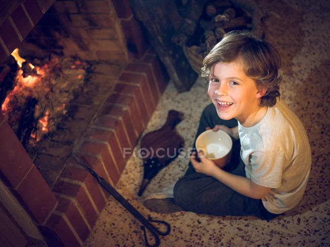 Мальчик сидит у камина с большой чашкой — стоковое фото