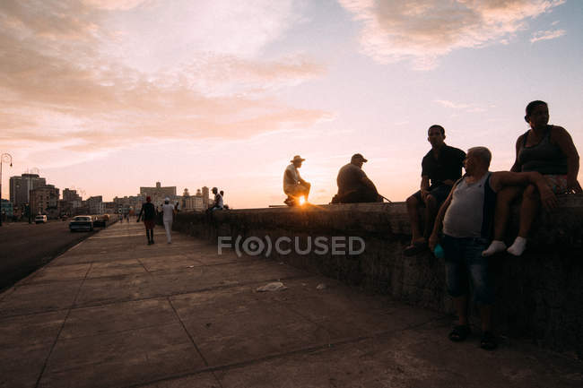 Cuban city dwellers chilling on pedestrian concrete waterfront on background at sunset — Stock Photo