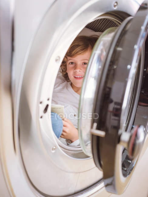 Menino da idade elementar deitado dentro da máquina de lavar roupa e livro de leitura . — Fotografia de Stock