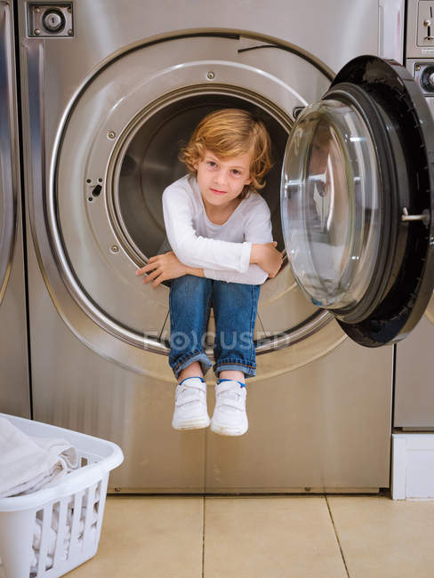 Bonito menino pré-escolar sentado dentro da máquina de lavar roupa e olhando na câmera . — Fotografia de Stock