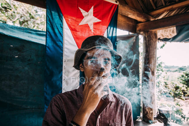LA HABANA, CUBA - MAY 1, 2018: Man in hat smoking thick cigar standing against flag of Cuba in small hut in sunlight — Stock Photo
