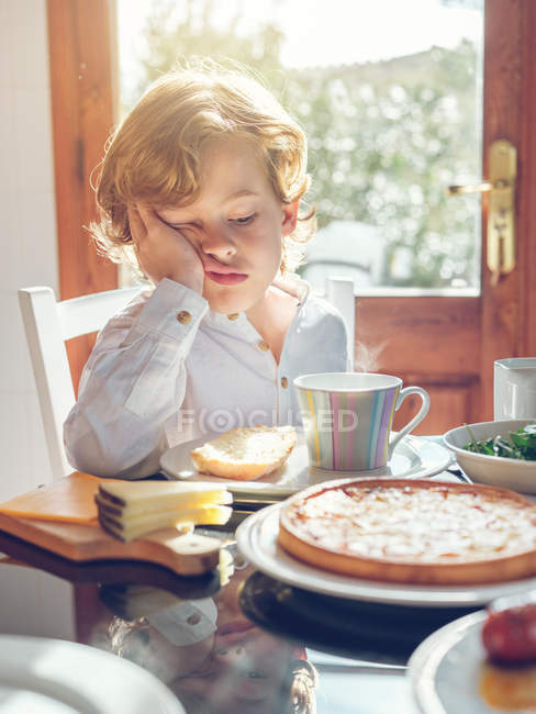 Annoiato ragazzo seduto con tazza a tavola — Foto stock