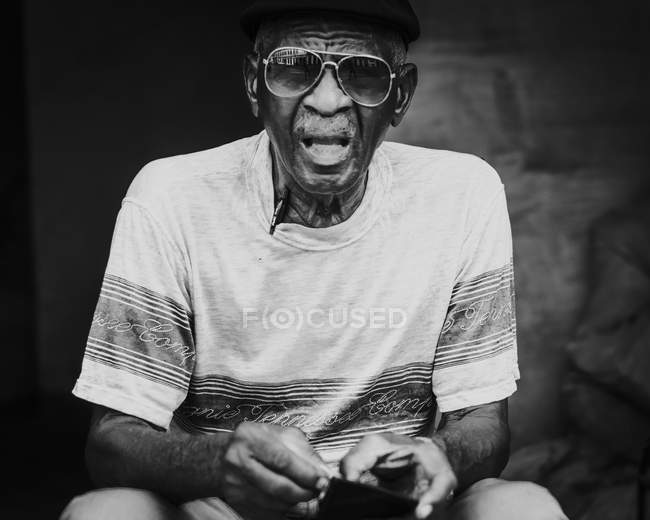 LA HABANA, CUBA - 1 de mayo de 2018: Anciano étnico con gafas de sol y camiseta con gorra sentado en la calle de Cuba - foto de stock