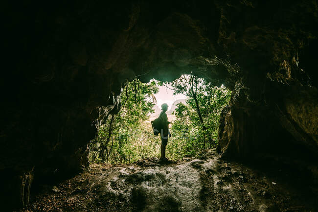 Vista misteriosa del viaggiatore con zaino in piedi sulla formazione rocciosa di ingresso grotta contro foresta verde tropicale. — Foto stock