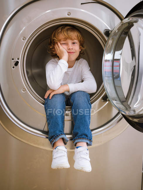 Elementary age boy sitting inside washing machine with hand on chin. — Stock Photo