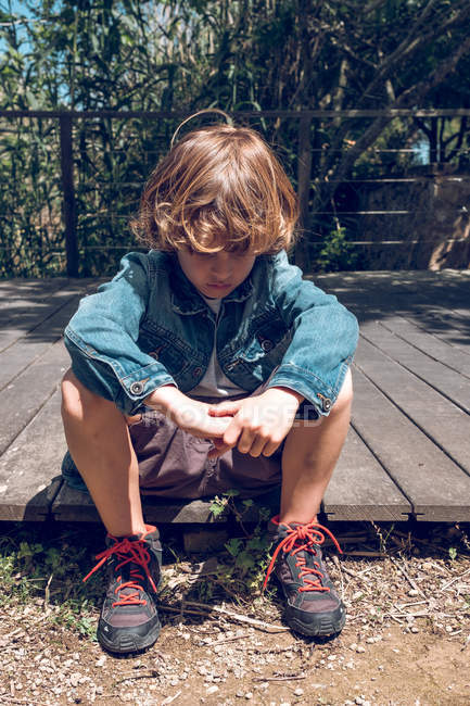 Cute elementary age boy with curly blond hair sitting on wooden bridge and looking down in countryside. — Stock Photo