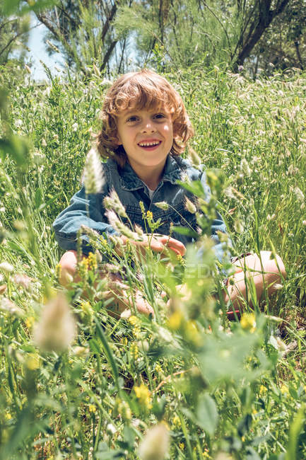 Menino da idade elementar sentado no campo de flores silvestres e sorrindo . — Fotografia de Stock