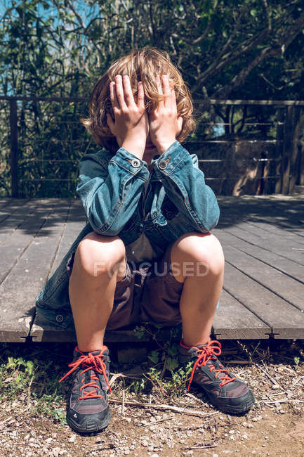 Elementary age boy with curly blond hair sitting on wooden bridge and hiding face in countryside. — Stock Photo