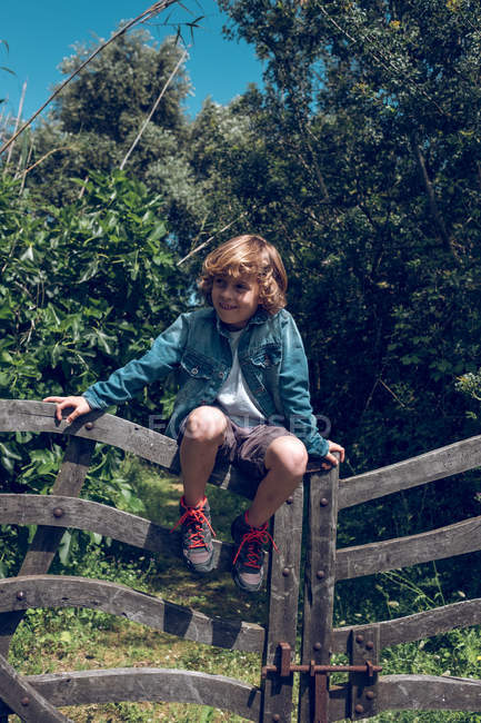 Junge im Grundalter mit blonden Locken sitzt auf einer Holzbrücke und lächelt im Grünen. — Stockfoto