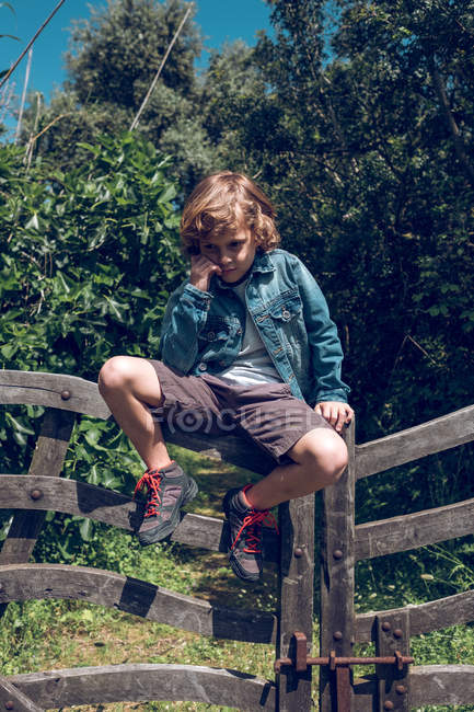 Junge im Grundschulalter mit blonden Locken sitzt auf Holzbrücke im Grünen. — Stockfoto