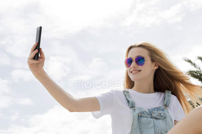 Menina loira sentada e tomando selfie na frente do céu nublado — Fotografia de Stock