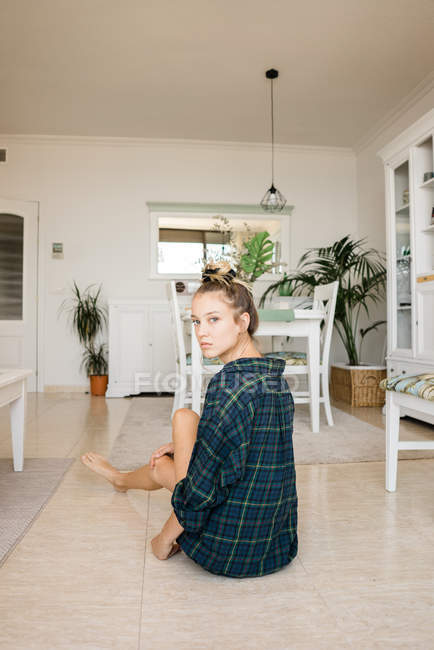 Молода жінка в картатій сорочці сидить на підлозі в стильній кімнаті і дивиться на камеру — стокове фото