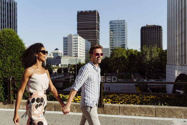 Щаслива багаторасова пара посміхається і тримає руки під час прогулянки по міській вулиці разом у сонячний день — стокове фото