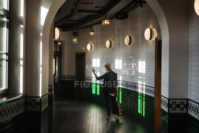 Visão traseira da mulher nova na moda usando telefone e tirar selfie no salão moderno de azulejos com lâmpadas — Fotografia de Stock