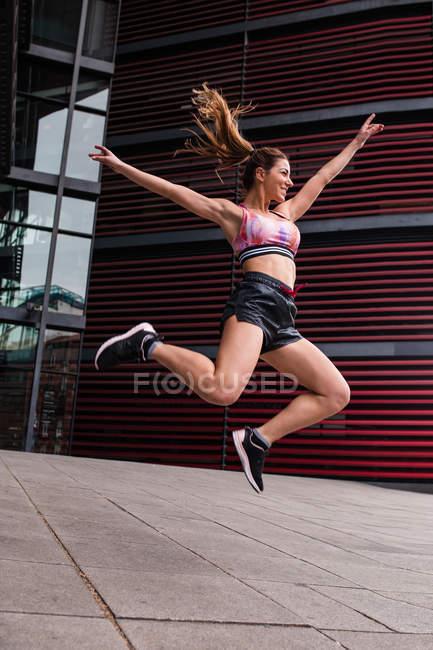 Femme en forme musculaire excitée en vêtements de sport sautant heureux avec les mains écartées sur la rue pavée — Photo de stock