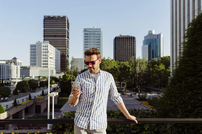 Елегантний чоловік в сонцезахисних окулярах спирається на паркан і використовує смартфон, перебуваючи в сучасному місті — стокове фото