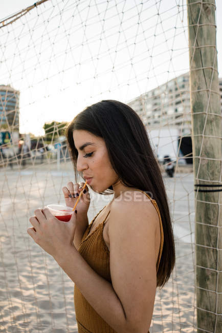 Молодая взрослая женщина с длинными волосами пьет лимонад на песчаном берегу — стоковое фото