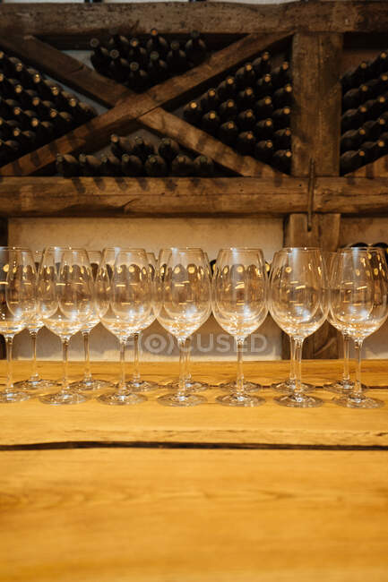 Fila de elegantes copas brillantes de pie sobre mesa de madera en bodega con botellas de vino en estantes sobre fondo - foto de stock