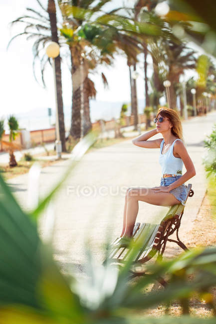 Женщина в летней одежде сидит на скамейке в тропическом парке — стоковое фото