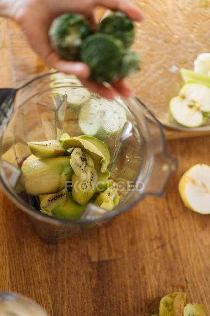 Main humaine mettant des ingrédients dans une tasse en plastique de mélangeur rempli de mélange de fruits et légumes pour smoothie — Photo de stock