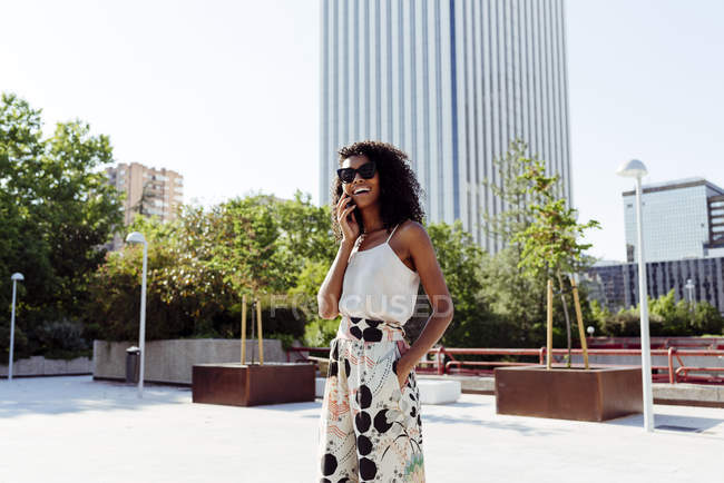 Элегантная афроамериканка, разговаривающая по смартфону, стоя на городской улице в солнечный день — стоковое фото