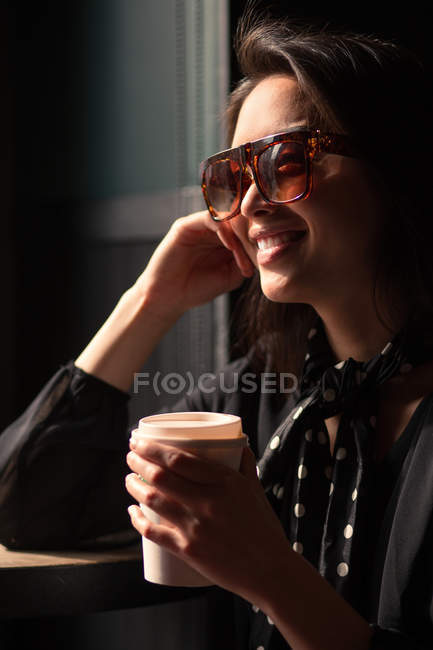 Модная элегантная улыбающаяся женщина в солнцезащитных очках и с платком, держа бумажную чашку кофе и опираясь на стол — стоковое фото
