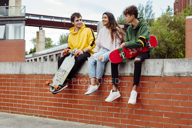 Alegres adolescentes multiétnicos masculinos y femeninos sentados con monopatines en una valla de ladrillo - foto de stock