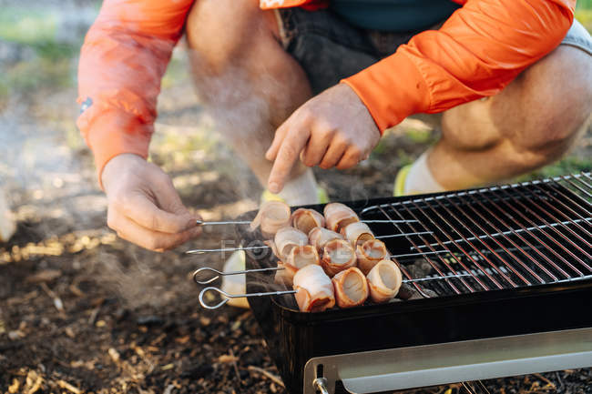 Male hands preparing bacon and sausages on skewers grilling on burning charcoal in portable griddle outdoors — Stock Photo