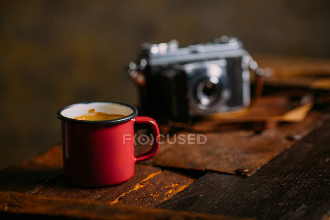 Enamel cup of coffee on rustic wooden surface with retro camera — Stock Photo