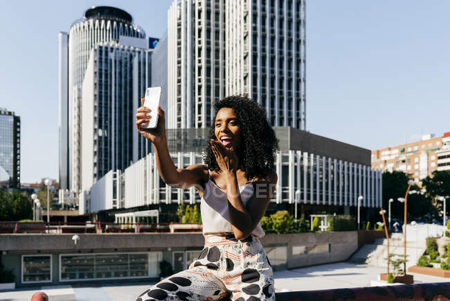Елегантний афро-американських жінка позує для selfie сидячи на паркан на міській вулиці на сонячний день — стокове фото