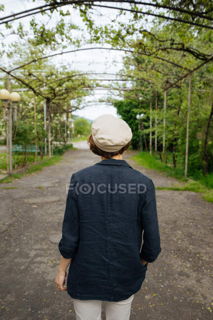 Back view of female in casual summer clothes and light beret walking on alley?under twined with vines arch corridor — Stock Photo