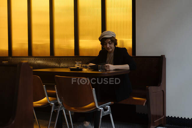Joven mujer bonita con gorra sentada sola en la mesa en el restaurante moderno y leyendo a través del menú - foto de stock