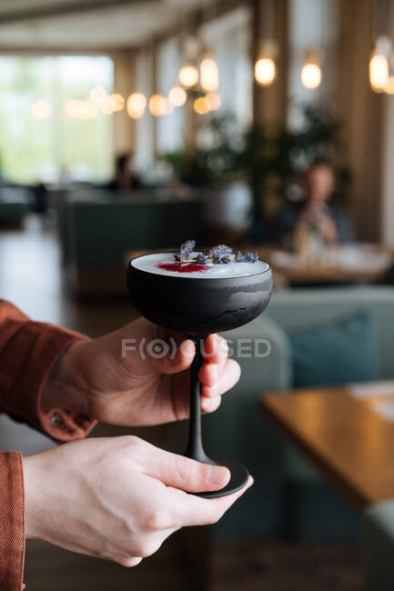 Crop view of male in orange shirt holding dark elegant stem glass with white Nordic beverage sprinkled with powder and dried flowers on blurred background — Stock Photo