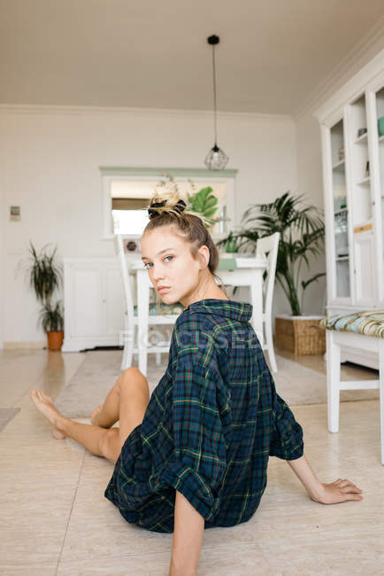 Молодая женщина в клетчатой рубашке сидит на полу в стильной комнате и смотрит в камеру — стоковое фото