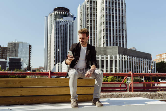 Елегантний хлопець переглядає смартфон сидячи на лавці на вулиці сучасного міста — стокове фото