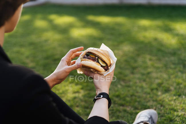 Mulher sentada na grama no parque e segurando takeaway hambúrguer — Fotografia de Stock