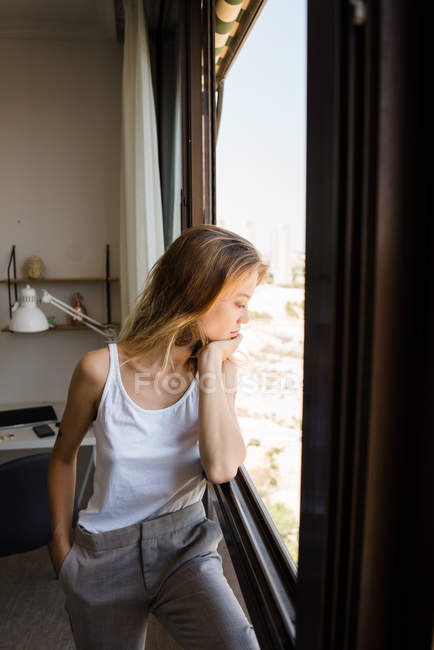 Nachdenkliche junge Frau steht und schaut mit dem Kopf auf der Hand durch das Fenster — Stockfoto