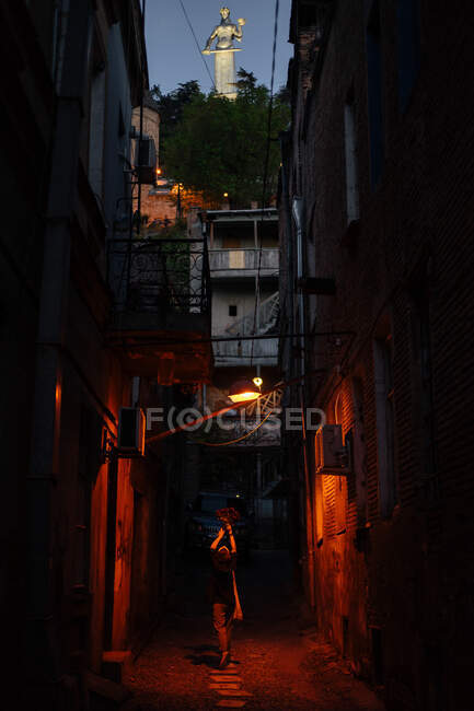 Back view of female walking in evening on narrow street illuminated by lamps holding bouquet in raised hands and looking up with marble statue of woman above — Stock Photo