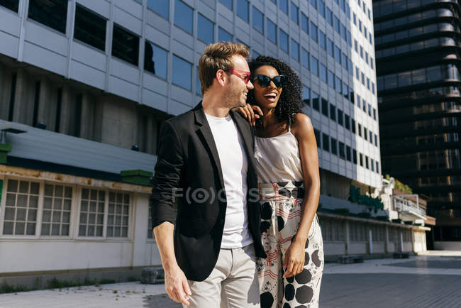 Элегантная многорасовая пара, идущая вместе по городской улице в солнечный день — стоковое фото