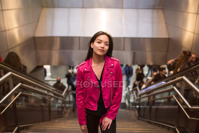 Портрет симпатичной брюнетки в розовой куртке, стоящей на ступеньках подполья и смотрящей в камеру — стоковое фото