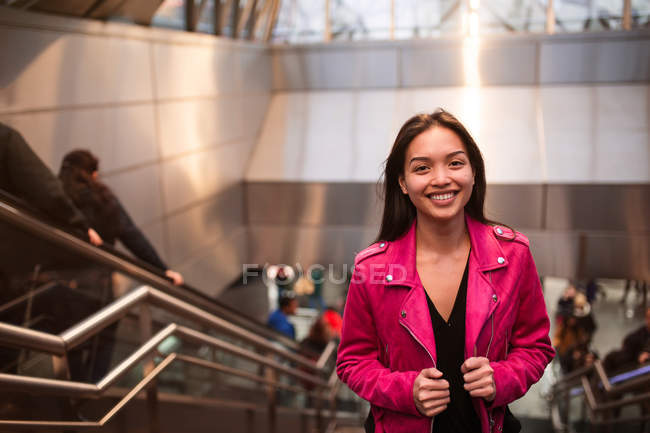 Portrait of pretty brunette in pink jacket standing on steps of underground and looking at camera — Stock Photo