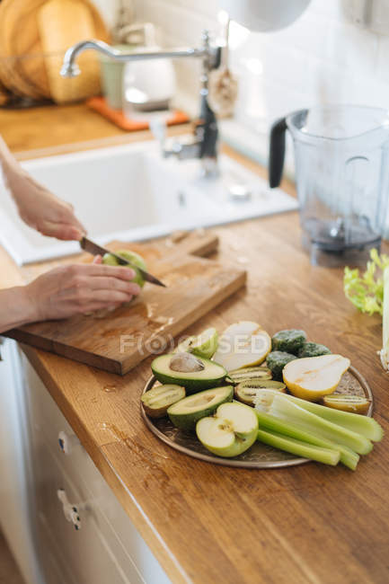 Mãos femininas cortando maçãs e preparando prato saudável com frutas e legumes verdes na superfície de madeira — Fotografia de Stock