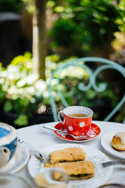 Red ceramic polka-dotted mug on saucer and pieces of pasty on plate on garden table — Stock Photo