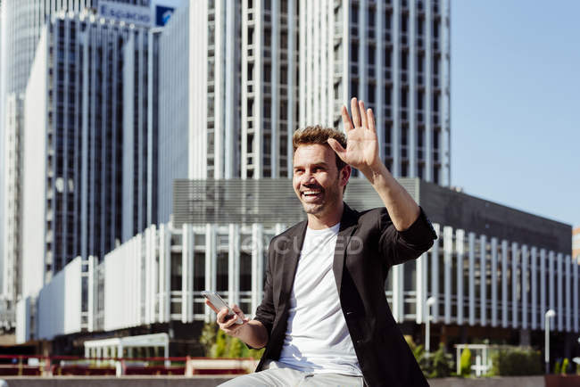 Man holding smartphone in modern city and waving hand — Stock Photo
