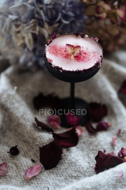Чудовий скандинавський напій у високому стовбуровому склі з червоним порошком і листом подається на сірій тканині з розсипаними пелюстками троянд на розмитому фоні. — стокове фото